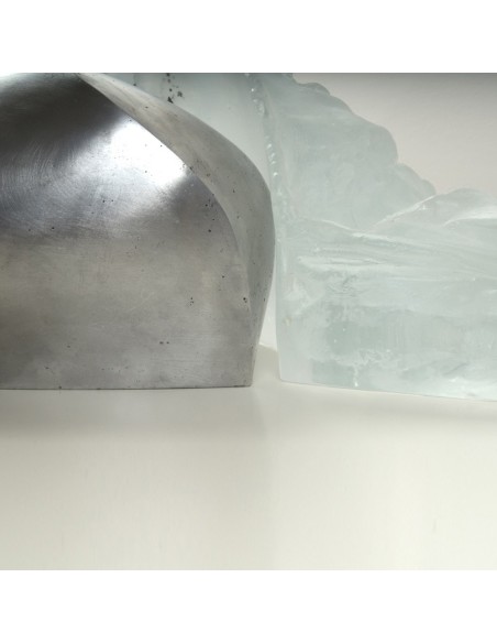 A close-up of a metallic, curved cremation urn beside a translucent ice-like sculpture on a white surface.