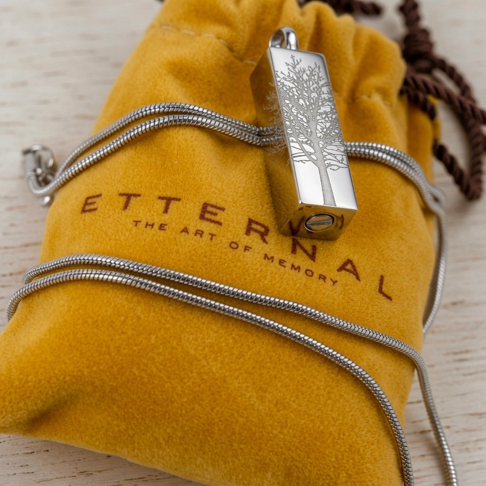 Cremation Jewell Tree Root Pendant on a silver chain rests on a yellow pouch with “ETERNAL” and “THE ART OF MEMORY” text visible