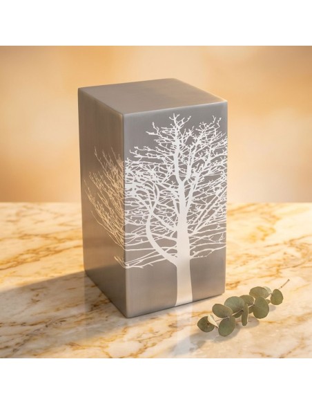 Cremation urn tree root design in a gray metallic box with white leafless tree illustration, on a marble surface.